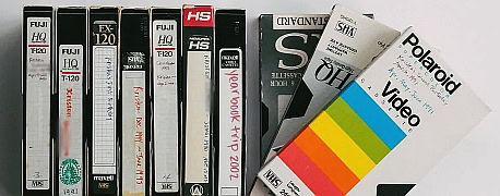 A row of VHS tapes in various cases stand upright, some labeled by hand with titles like yearbook trip 2002, next to several stacked and slanted videotape boxes, including a colorful Polaroid Video box.