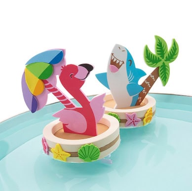 Two colorful drink floats shaped like a pink flamingo with an umbrella and a smiling blue shark with a palm tree, decorated with starfish and seashells, sit on a light blue surface.