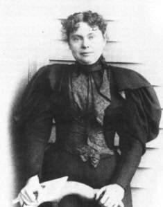 Black-and-white photo of a woman with curly hair wearing a high-necked, dark Victorian-era dress with puffed sleeves, standing in front of a wooden wall and holding an object.