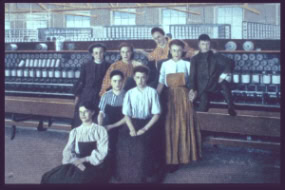 Eight women pose in front of a large textile machine inside a factory. They wear long skirts and blouses typical of the early 1900s. Rows of spools of thread are visible on the machine behind them.